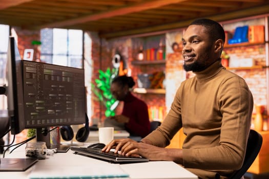 Happy man working in AI tech agency providing custom software development services. Cheerful african american IT expert using machine learning to streamline coding process in startup office