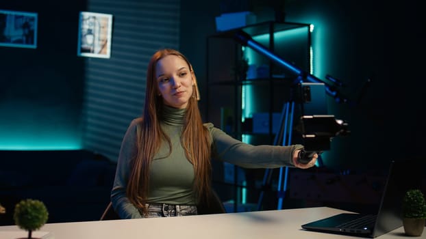 Upbeat teenager films content for online streaming channel in apartment studio using phone on selfie stick. Smiling gen z influencer at home interacts with subscribers, recording with phone, camera B