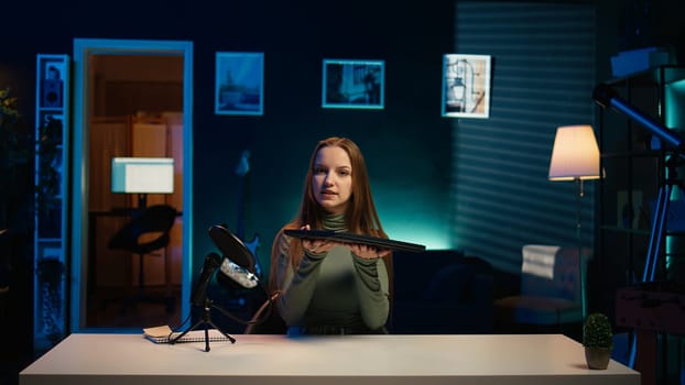 Young entertainer presenting sleek laptop from brand sponsoring video, urging viewers to purchase it. Girl does influencer marketing by advertising thin notebook to audience in studio, camera B
