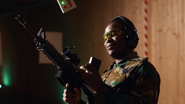 Portrait of cheerful soldier doing training in shooting range, reloading rifle. Joyous military unit in firing range practicing with semiautomatic weapons, wearing protective equipment, camera B