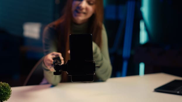 Close up of phone on selfie stick used by girl in apartment to capture intro for vlogging channel. Focus on smartphone used by entertainer in blurry background to film during live stream, camera A