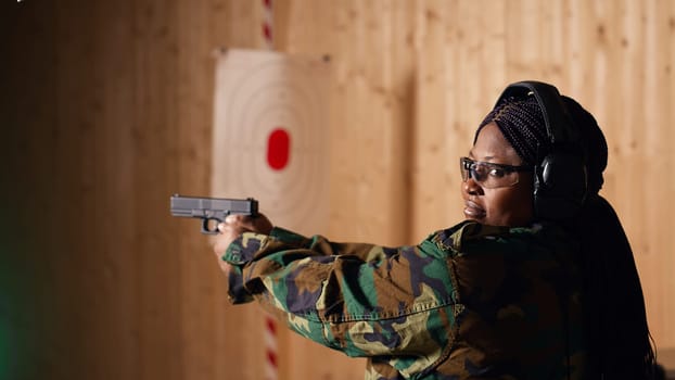 Portrait of smiling woman in firing range shooting with pistol, wearing earmuffs to protect hearing. Cheerful person in shooting gallery using safety gear to prevent damage from gunshots, camera B