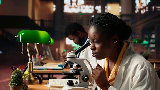 Female science student examines cells under the microscope for her scientific research in a campus library, takes notes about chemical processes and reactions for her thesis. Camera A.