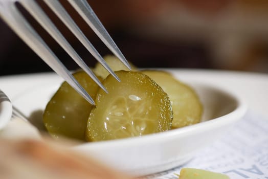 A fork lifts a slice of pickle from a dish containing more pickles.