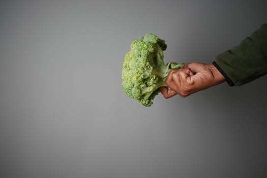 A hand grips a fresh, vibrant broccoli against a neutral gray backdrop.