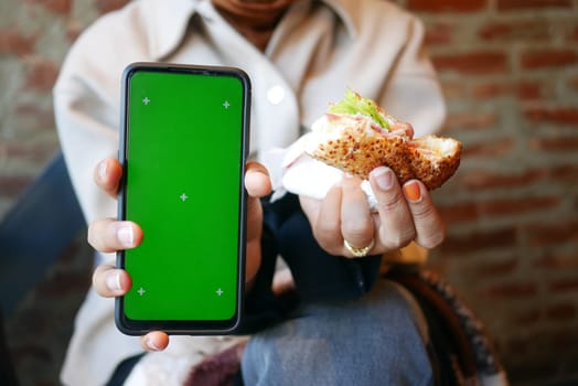 A person holds a sandwich and a smartphone with a green screen.
