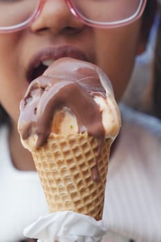 A young child bites into a melting chocolate ice cream cone outdoors.