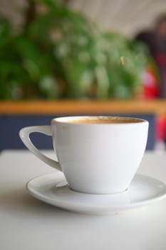 A white coffee cup is placed on a table near lush green plants.