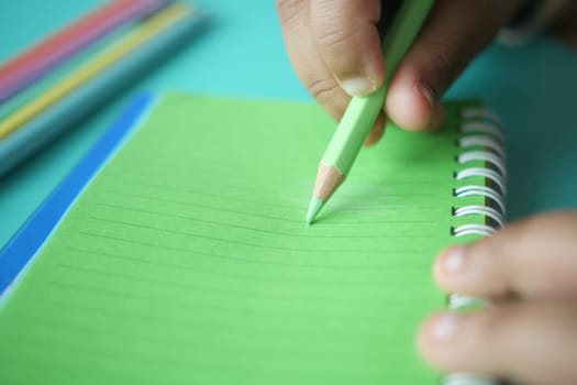 A hand holding a green pencil makes notes on a lined notebook.