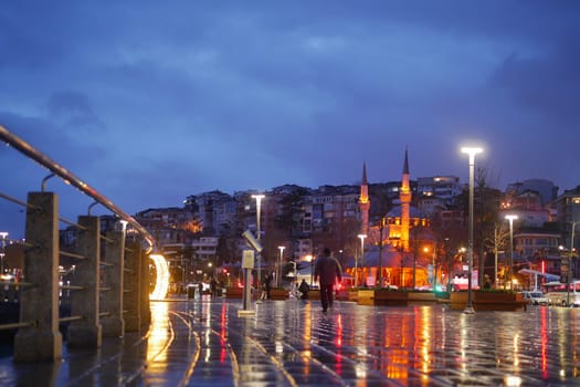 People stroll by the waterfront as lights reflect on the wet pavement.