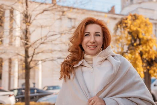 Smiling redhead woman in a beige coat and shawl stands outdoors, before a pale stone building and bare trees. Autumnal setting.