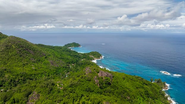 Aerial view of a lush green coastline with hills meeting the blue ocean under a cloudy sky. Seychelles, Mahe.