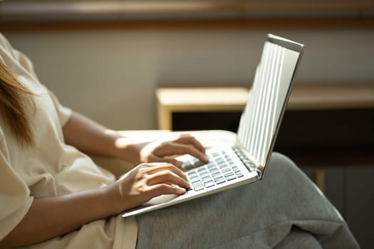 Close up of woman typing on a laptop while sitting comfortably in a sunlit room.