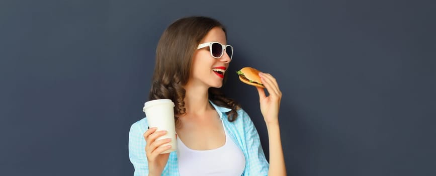 Portrait of happy cheerful young woman eating burger fast food holding cup of coffee, teenage girl having fun isolated on black wall studio background