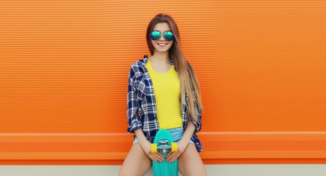 Summer portrait of happy cheerful stylish young woman with skateboard, teenager girl in colorful clothes, glasses posing on orange background on city street
