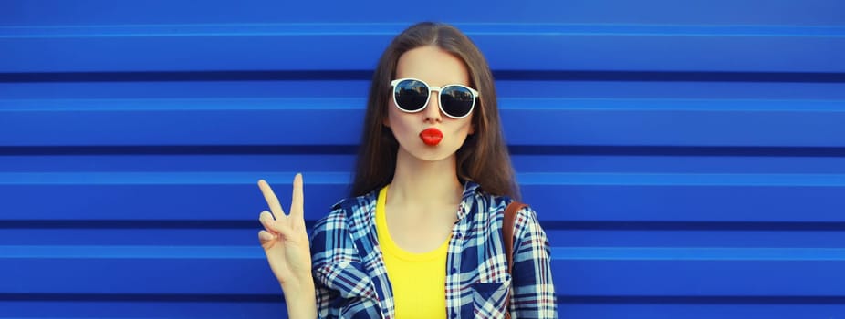 Portrait of stylish young woman blowing a kiss, cool modern girl posing in casual clothing on blue wall background