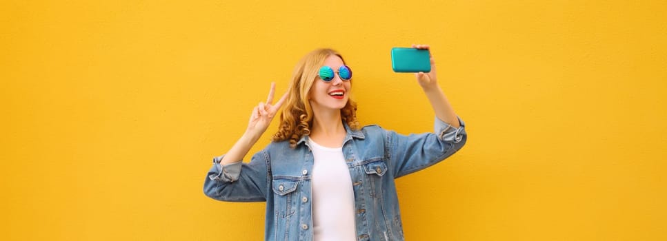 Joyful happy young woman laughs taking selfie with phone, modern cheerful girl posing in casual denim clothing on bright yellow background