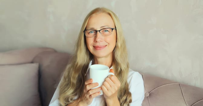 Happy satisfied mature woman enjoying hot tasty cup of coffee or tea looking at camera in early morning sitting on a couch in the living room at home