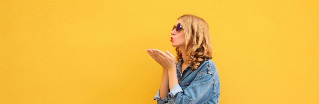 Beautiful stylish young woman blowing kiss in glasses, denim jacket posing on yellow wall background, blank copy space