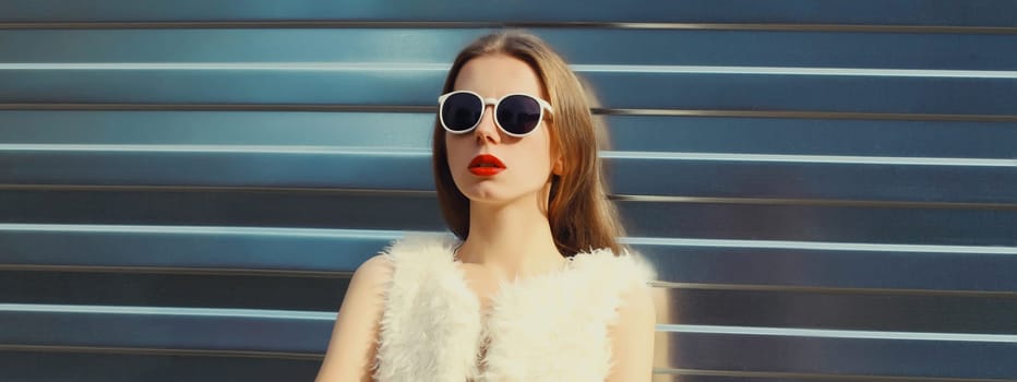 Portrait of modern stylish young woman in white fur vest, glasses, fashionable female model posing on gray silver wall background