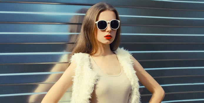 Portrait of modern stylish young woman in white fur vest, glasses, fashionable female model posing on gray silver wall background