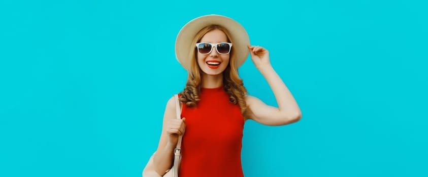 Portrait of beautiful happy young woman in summer tourist straw hat, red dress and glasses, pretty girl smiles with backpack posing on bright blue background