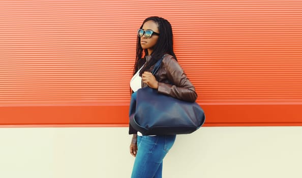 Fashionable young African woman with handbag in casual clothing, stylish modern black girl with dreadlocks, glasses walking in the city on red background
