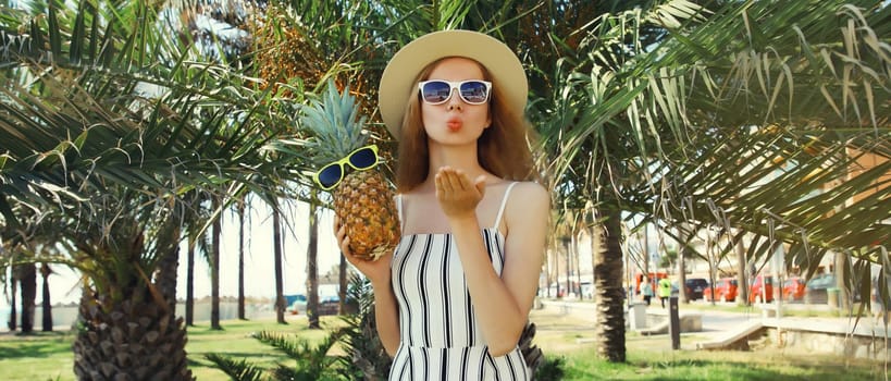 Summer fashion outfit of trendy young woman blowing a kiss holding pineapple fruits wearing hat, striped clothes, sunglasses posing in park with palm trees background