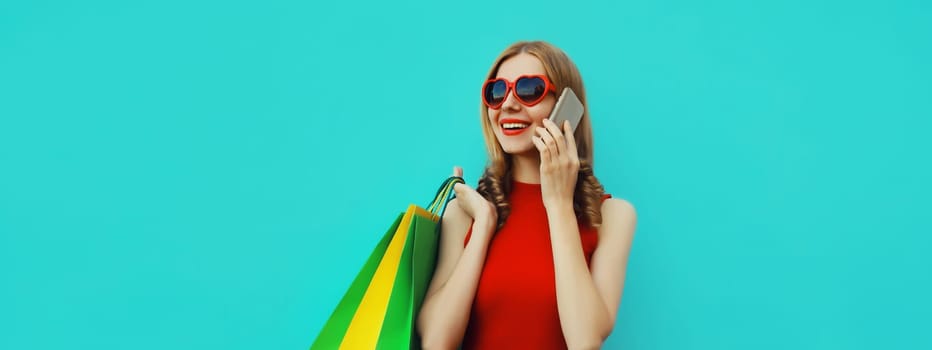 Beautiful happy young woman calling on smartphone with shopping bag, pretty girl smiles looking away in red dress, heart shaped glasses standing on blue background