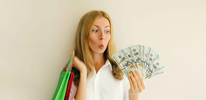 Portrait of surprised shocked young woman with shopping bags holding cash money looking at dollar bills on white background