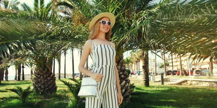 Summer outfit, stylish beautiful young woman posing in white striped jumpsuit with handbag standing in green park with palm trees background