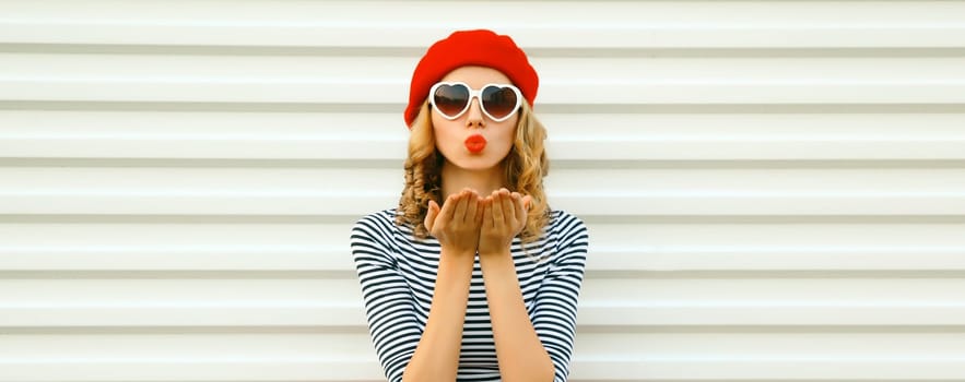 Portrait of beautiful happy young woman blowing a kiss with lipstick in red french beret, heart shaped sunglasses on white background