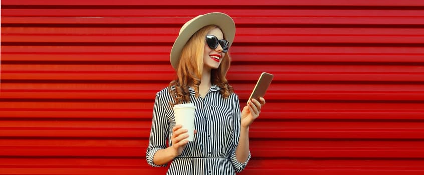 Summer portrait of stylish happy young woman with mobile phone looking at device in straw hat, holding cup of coffee on red background