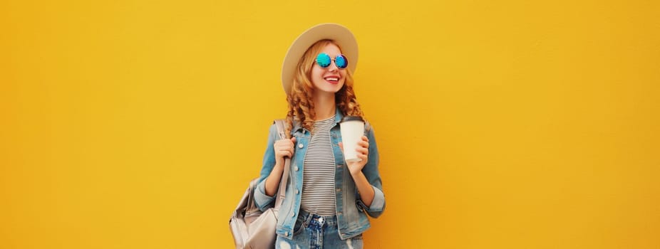 Beautiful stylish happy smiling young woman with cup of coffee wearing summer hat, backpack, jean jacket on yellow background