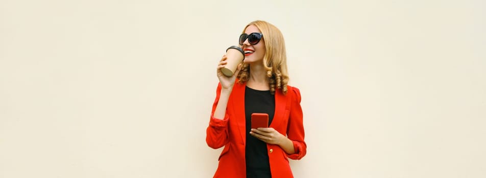 Stylish happy young businesswoman drinking coffee with phone, beautiful woman using smartphone holding cup of coffee dressed red business suit looking away standing on white background