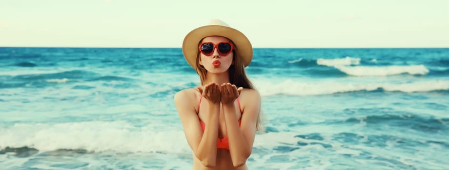 Summer vacation, beautiful happy lovely woman blowing a kiss in pink bikini swimsuit, tourist straw hat, red heart shaped glasses on the beach on sea coast with waves