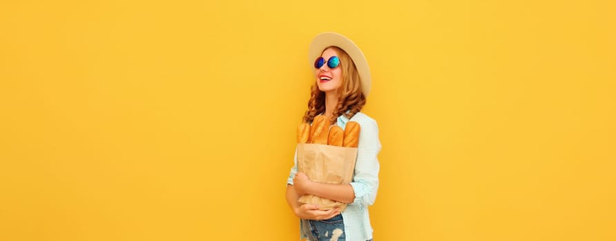 Happy cheerful young woman holding grocery shopping paper bag with fresh long white bread baguette looking away on yellow studio background