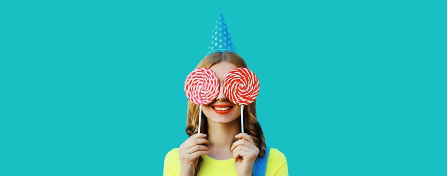 Holiday, celebration, event. Happy cheerful young woman laughs celebrating having fun at a party wearing festive birthday hat holding lollipop on blue background
