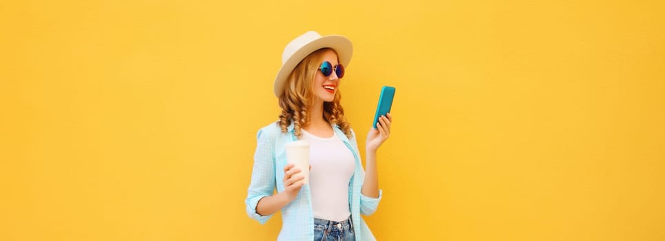 Summer stylish happy young woman with phone, joyful girl looking at smartphone holding cup of coffee dressed tourist straw hat standing on yellow background