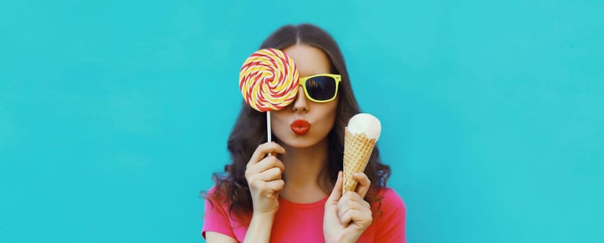 Summer portrait of happy young woman holding ice cream cone and lollipop blowing a kiss on blue background