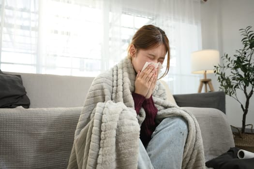 Young woman suffering from flu symptoms wrapped in blanket, sneezing into a tissue..