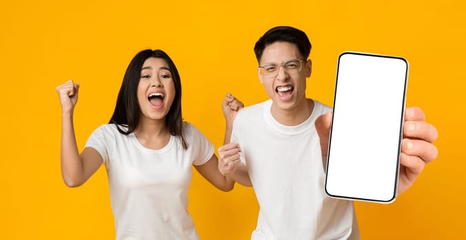 Yes. Excited Asian Couple Holding Big Smartphone With Empty White Cell Screen In Hand Shaking Clenched Fists, Cheerful Guy And Lady Celebrating Win, Yellow Orange Studio Background, Mock Up Copy Space