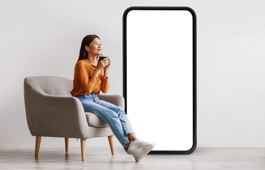 Young Asian woman drinking coffee, having break, relaxing in armchair near giant cellphone with empty screen against white studio wall, mockup for cool new mobile app or online ad design