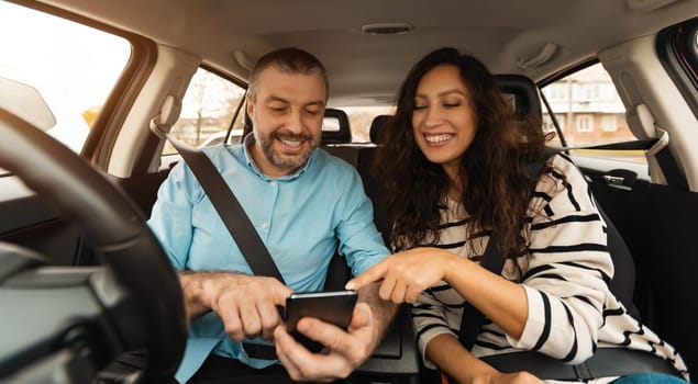 Short Road Route. Portrait of smiling couple sitting inside luxury car pointing fingers at smart phone, cheerful male driver holding cell, showing choosing location, using digital map application