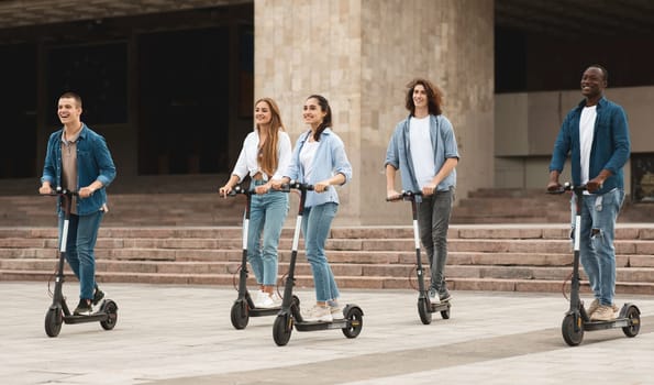 Carefree Youth. Full body length shot of five multicultural casual friends riding electric kick scooters in the city centre, spending free time together on weekend or vacation outdoors