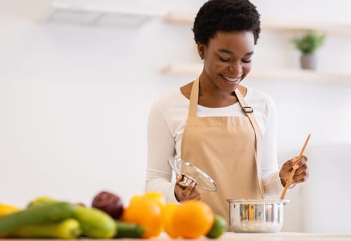 Happy African Woman Cooking Dinner In A Pot Standing In Modern Kitchen At Home, Wearing Apron. Black Housewife Preparing Meal Indoors. Healthy Food Recipes, Household And Nutrition