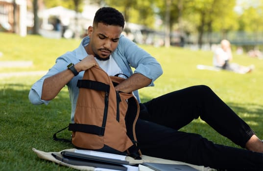 Serious african american guy looking inside his backpack and searching something, resting in city park outdoors. Black male student getting something out of his bag