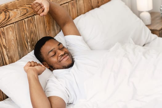 Good Morning. Rested Black Man Stretching Hands Waking Up Lying In Comfortable Bed At Home, Smiling With Eyes Closed. Wellslept Man Awakening After Good Sleep And Rest. High-Angle View