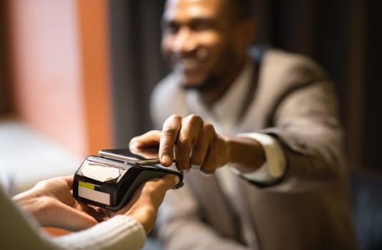Contactless Payment System. Afro guy in suit holding smartphone close to electronic payment machine, paying for delivery