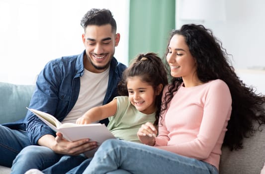 Family Time. Happy Arab Mom, Dad And Little Daughter Reading Book Together While Relaxing On Couch In Living Room, Happy Middle Eastern Parents Having Fun With Their Child At Home, Closeup Shot
